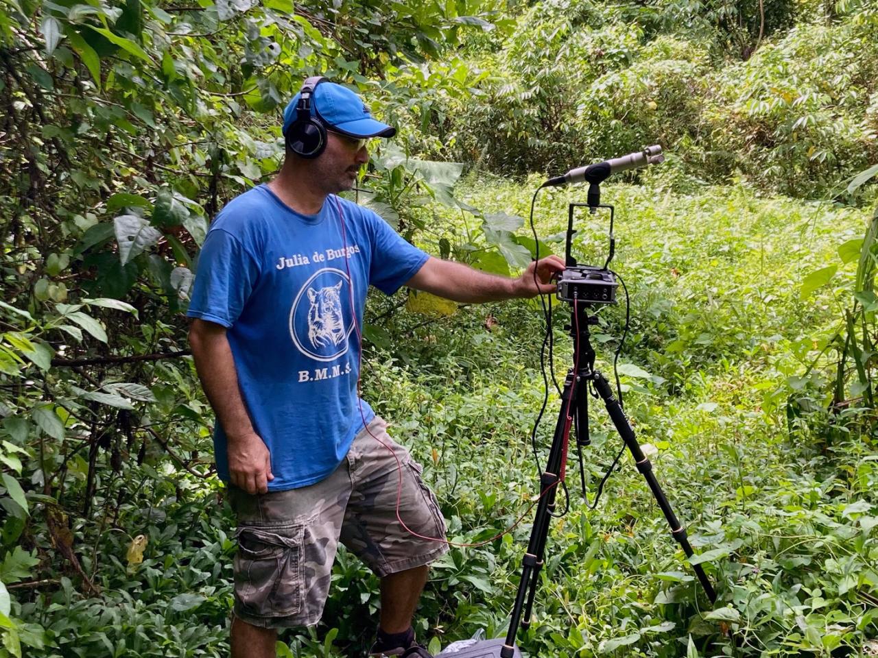 Filmmaker Juan Manuel Pagán Teitelbaum stands next to a recording device which is placed on a tripod. He is surrounded by lush greenery.